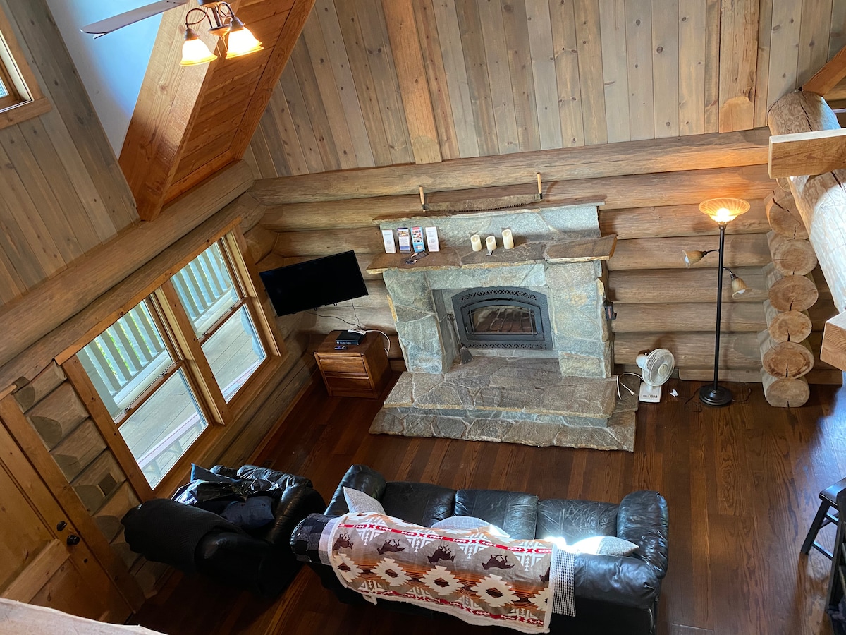 The living area showcases a stone fireplace as its focal point, surrounded by wooden walls and polished flooring. Two leather recliners are positioned for comfort, and a television is mounted nearby. Natural light enters through large windows, enhancing the cozy ambiance of the space.