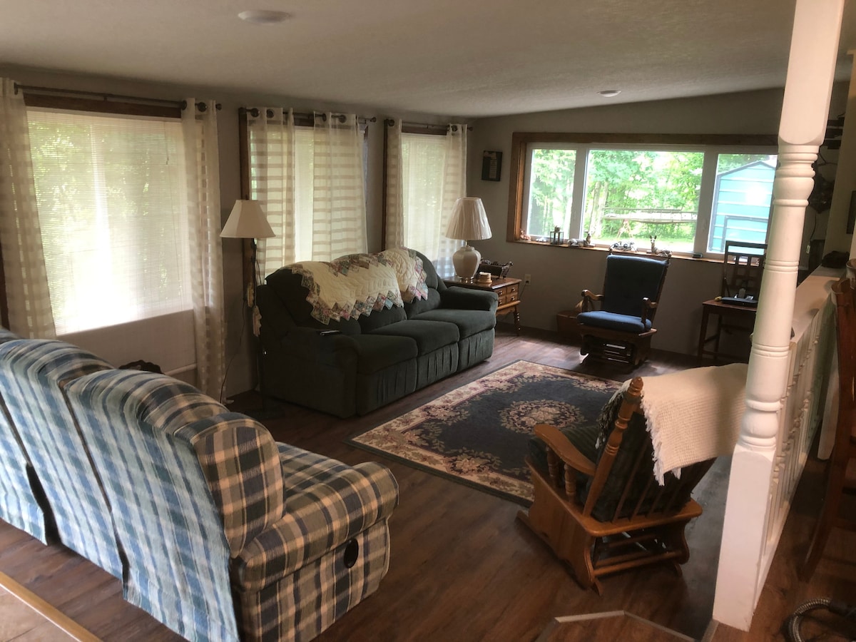 A spacious living area features comfortable seating arrangements, including a green couch and a plaid-patterned loveseat. Two lamps provide soft lighting. Natural light floods in through several windows, highlighting a decorative area rug. A rocking chair and a small table add to the inviting atmosphere.
