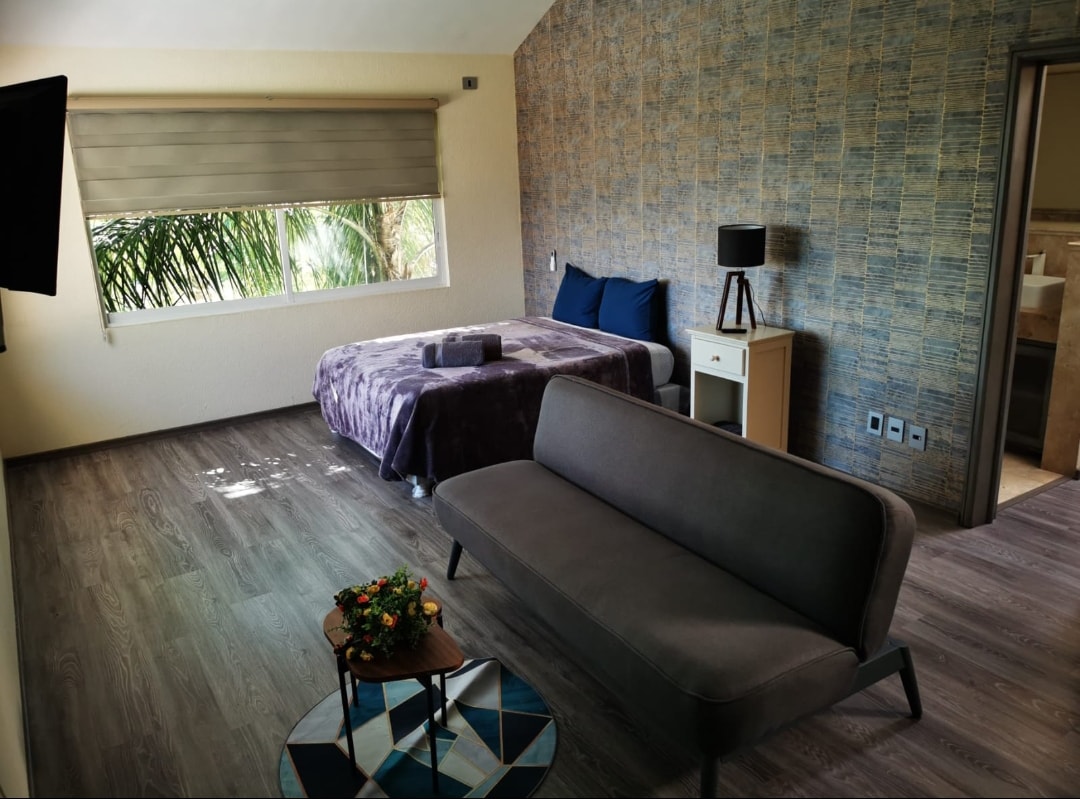 A spacious bedroom features a large bed covered with a muted throw. A comfortable sofa sits beside a small coffee table, and a window offers views of greenery outside. Textured wall accents create a soothing backdrop.
