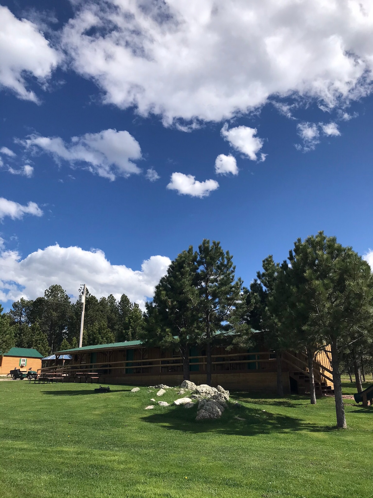 A spacious outdoor area is visible, featuring a long wooden building with multiple points of entry. Pine trees are positioned around the area, and a cloudy blue sky creates a scenic backdrop. Rocky outcrops are scattered across the manicured green lawn.