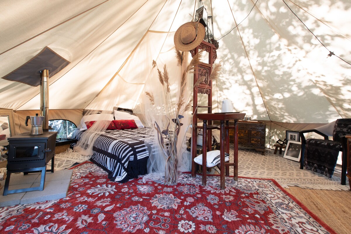 An interior view of a canvas bell tent displays a queen bed with black and white bedding, surrounded by decorative rugs. A wood-burning stove is positioned nearby, complemented by a small table and chairs, while natural light filters through the fabric walls, enhancing the overall ambiance.