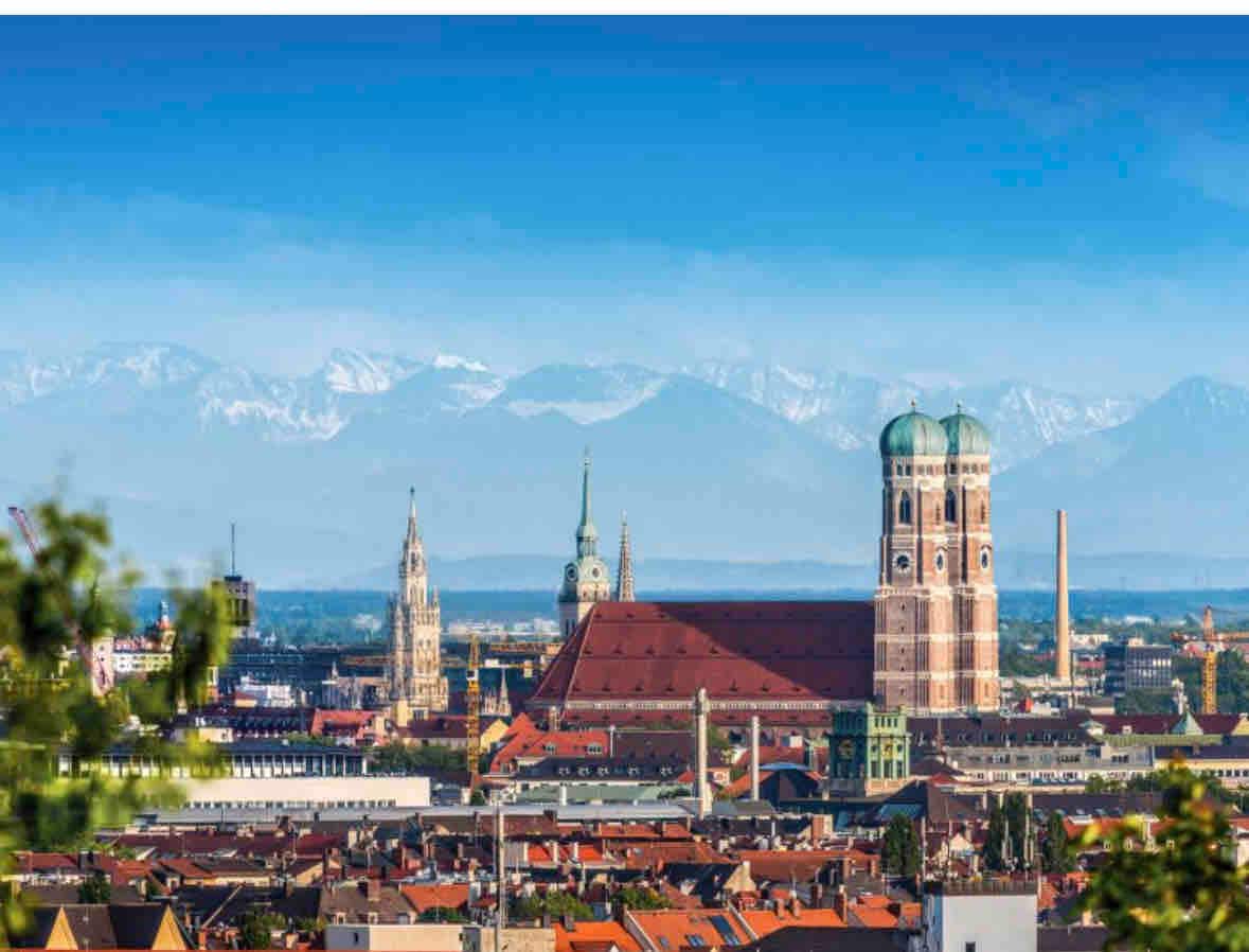 A panoramic view of the city skyline highlights historic architecture alongside modern structures. The prominent twin towers of a cathedral rise over rooftop landscapes, with snow-capped mountains visible in the background, creating a vast and scenic cityscape under a clear blue sky.