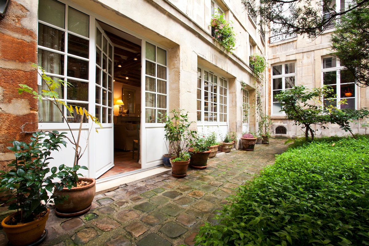 A peaceful courtyard is framed by the elegant façade of the building, featuring multiple large windows with white frames. Potted plants line the stone walkway, leading to a lush green garden area, enhancing the serene ambiance of this outdoor space.