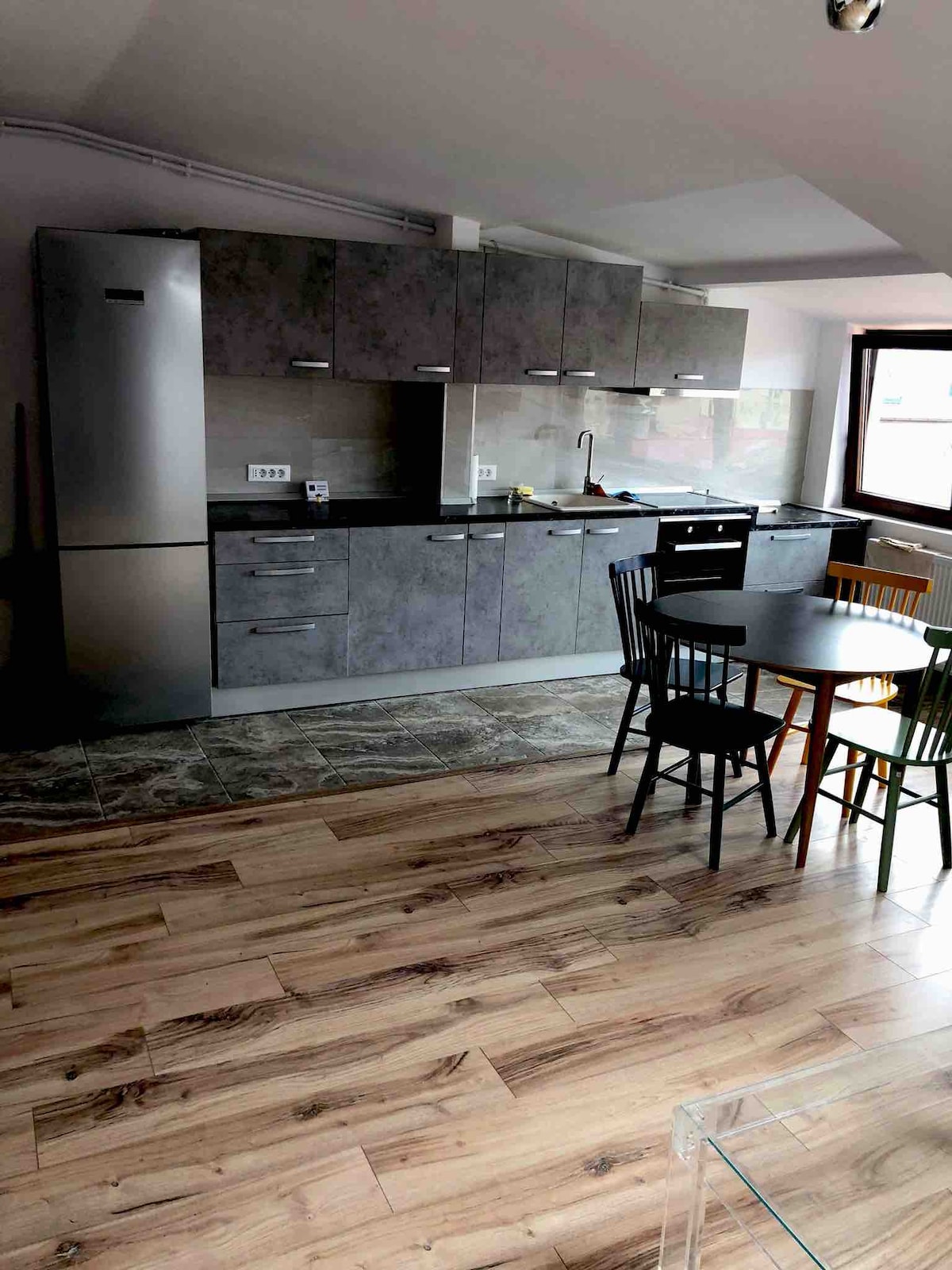 The kitchen is equipped with modern appliances and sleek cabinetry. A dining table made of wood is accompanied by a mix of black and natural wood chairs. The flooring features wooden planks, contributing to a contemporary feel, while natural light enters through a window.