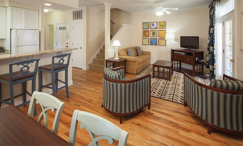 An open living area features a comfortable sofa and a coffee table, complemented by two striped armchairs. A decorative area rug enhances the space, with a dining table visible. A television is placed on a wooden console, while light streams in through the glass doors.