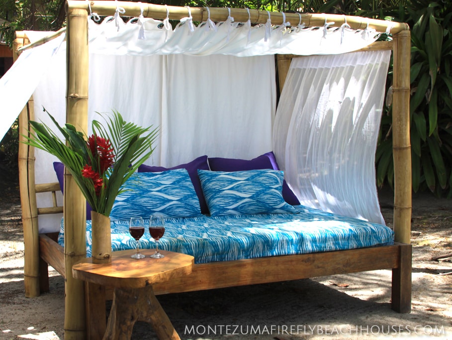 A bamboo chill-out bed is surrounded by flowing white fabric, creating a relaxing retreat. Soft blue bedding features a wave pattern on a spacious mattress. Nearby, a small wooden table holds two glasses of dark beverage and a vibrant tropical leaf arrangement, inviting leisurely moments in the garden.