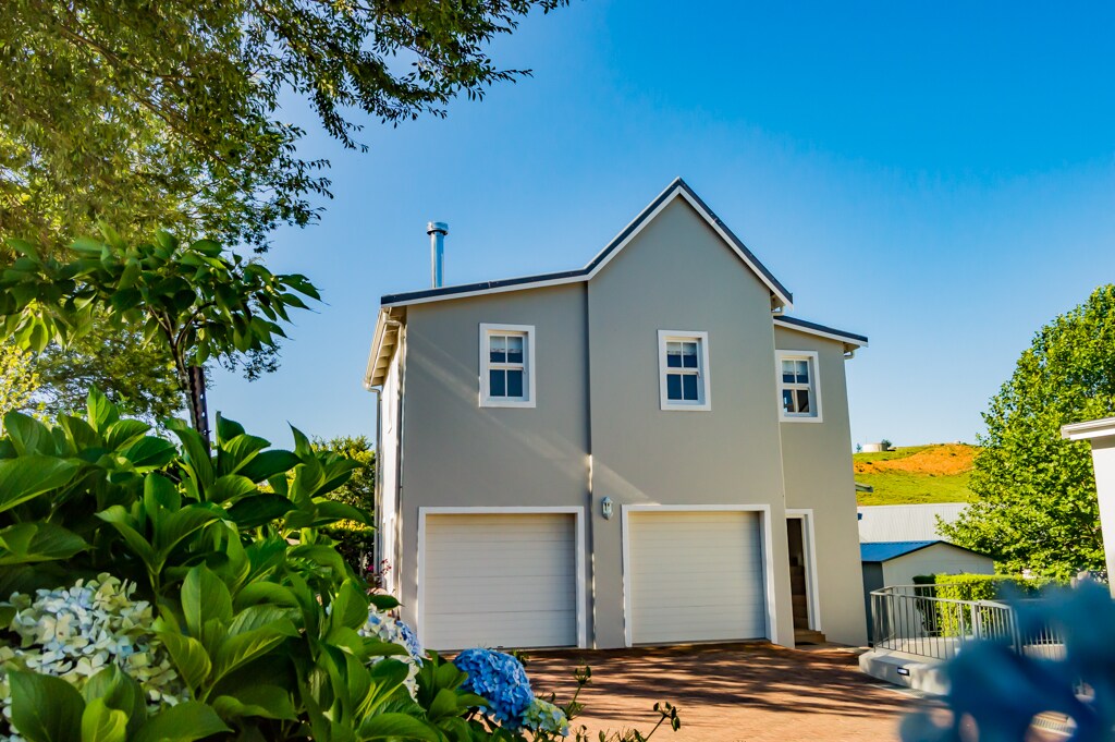 The exterior features a two-story structure with a light grey facade, highlighted by large white garage doors. Lush greenery and colorful flowers frame the entrance, while a clear blue sky adds a bright backdrop. The overall setting conveys a welcoming atmosphere.
