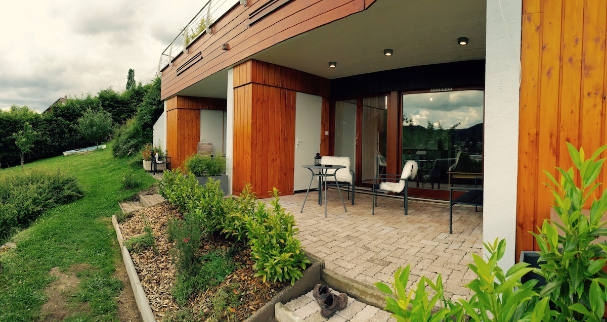 A spacious private loggia features a table with chairs, ideal for outdoor relaxation. Surrounding greenery adds a natural touch, while the stone-paved area provides ample space. The structure showcases a harmonious blend of wood and modern design, with large windows inviting natural light.