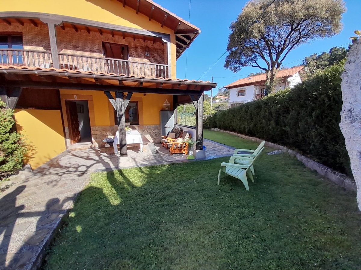 A charming outdoor area features a well-kept lawn, bordered by tidy hedges. Comfortable seating is arranged on a patio covered by a wooden overhang. A sunlit space allows for relaxation, accentuated by a tree providing shade nearby.