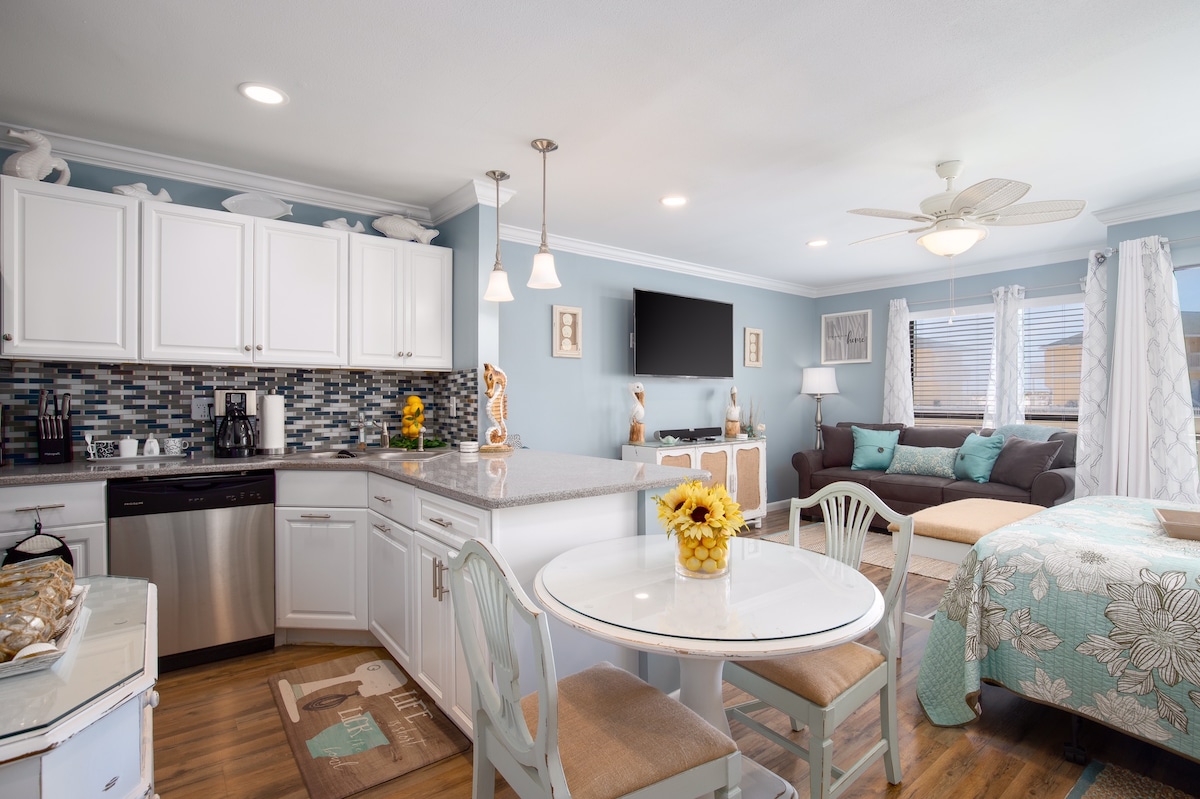 A well-appointed studio condo features a modern kitchen with a stainless steel refrigerator and ample counter space. A round dining table with four chairs sits beside a cozy sitting area that includes a sleeper sofa and a colorful quilted bed, all bathed in soft, natural light.