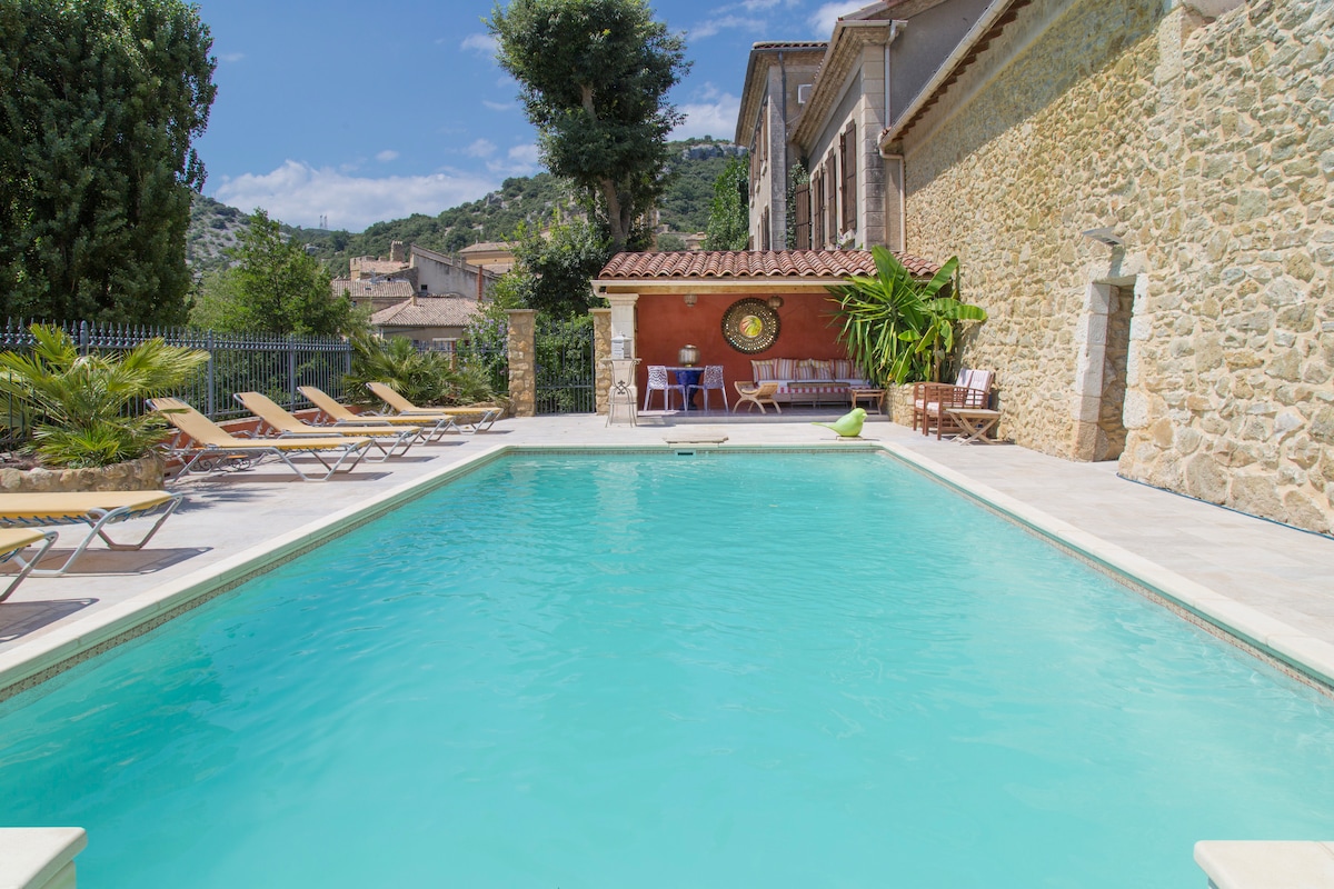 A clear swimming pool is framed by a sunbathing area featuring several lounge chairs. The pool's turquoise water reflects the blue sky above, while a stone building and lush greenery are visible in the background, providing a serene setting.