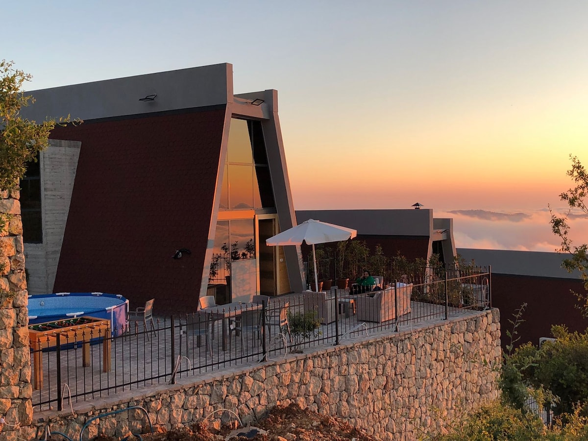 A modern bungalow is set on a hillside, featuring a stone terrace with seating and an umbrella. A blue pool is positioned nearby, framed by lush greenery. Large glass windows reflect the colors of the sunset sky, creating a serene atmosphere.