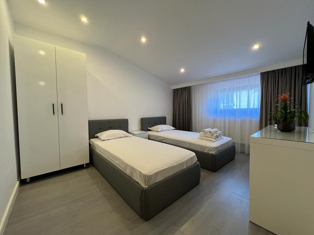 A bright sleeping area is featured with two beds, each equipped with white linens. A closet stands against one wall, while a sleek dresser is positioned nearby. Natural light filters through a sheer curtain, illuminating the space.