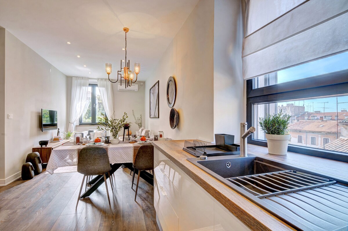 The dining area features a modern table set for breakfast, complemented by stylish chairs. A sleek kitchen sink is visible, with a window above that allows natural light to pour in. Decorative plants are placed on the windowsill, enhancing the inviting ambiance.