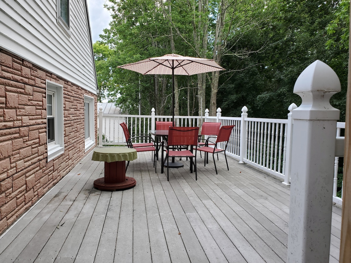 An outdoor deck is visible, featuring a round table surrounded by six red chairs and a large umbrella for shade. A circular planter is positioned nearby on the wooden flooring, which shows a neutral tone. Trees in the background provide a natural setting.