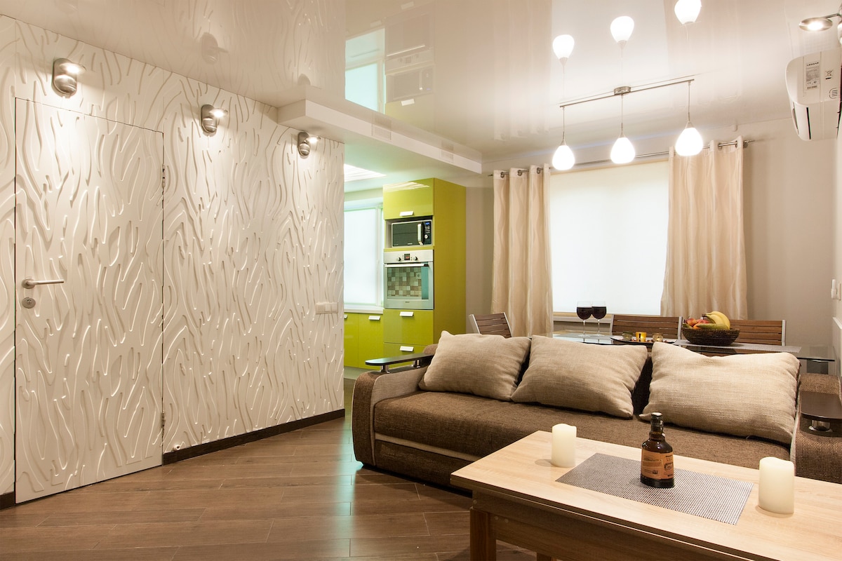 A spacious living area features a comfortable sectional sofa with soft pillows facing a wooden coffee table. Light fixtures hang from the ceiling, and natural light enters through large windows. A kitchen area is visible in the background, with modern appliances and a dining table.