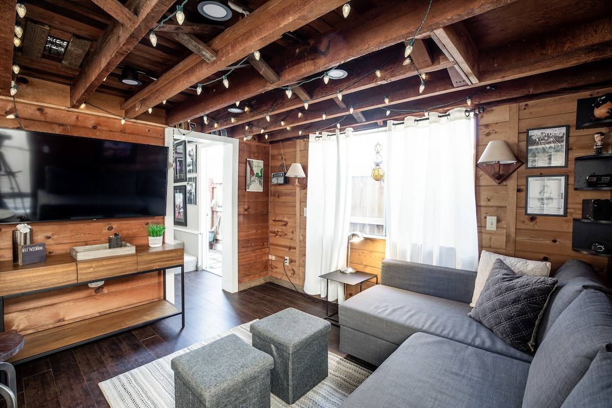 The living area features wooden beams and walls, creating a rustic ambiance. A large sectional couch is positioned next to a low table, with two ottomans nearby. Natural light enters through the white sheer curtains, complementing the mounted television and decorative wall art.