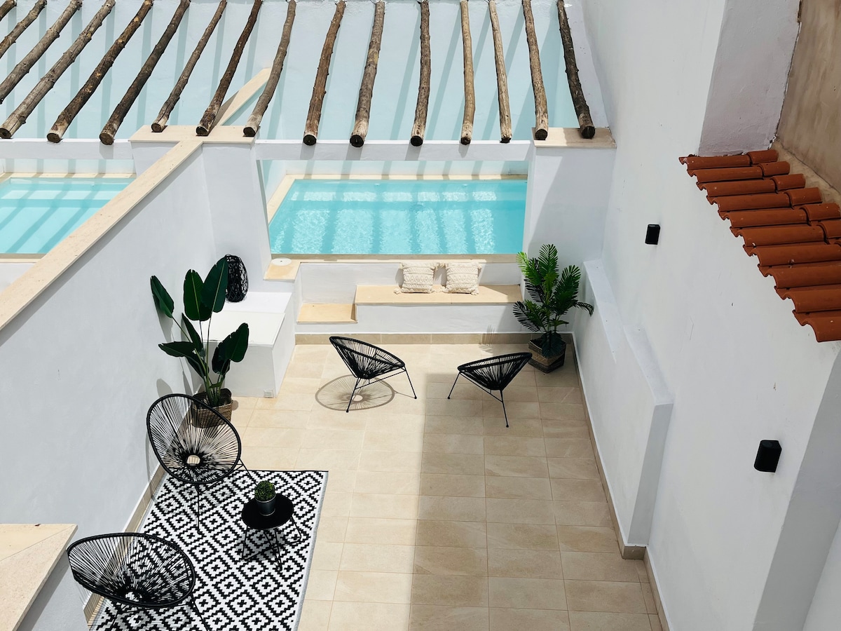 An outdoor area features a private pool surrounded by light-colored tiles. Two black lounge chairs are placed on a patterned rug, while potted plants add greenery to the space. Natural light filters through wooden beams overhead, enhancing the serene atmosphere.