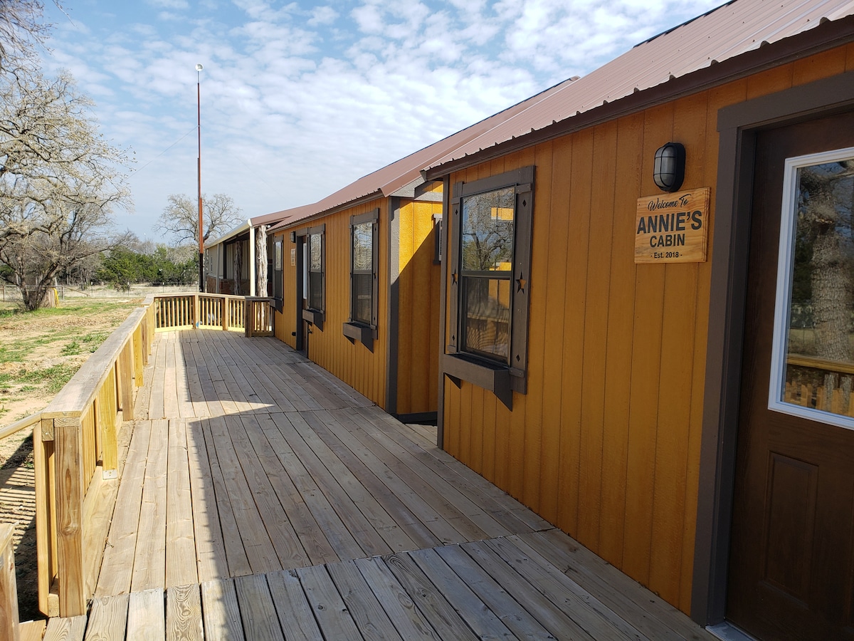 A wooden deck extends along the cabin's exterior, bordered by bright yellow siding. Multiple windows allow natural light to fill the interior. A sign identifying 'Annie's Cabin' is mounted next to the door, and open space with rural scenery is visible in the background.