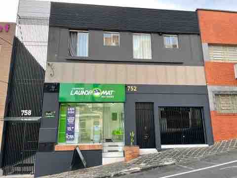 The building exterior features a modern design with a dark façade and a prominent green sign for Laundromat. A well-defined entrance is visible, and the structure is nestled among neighboring buildings. The sloped street in front adds to the unique urban environment.