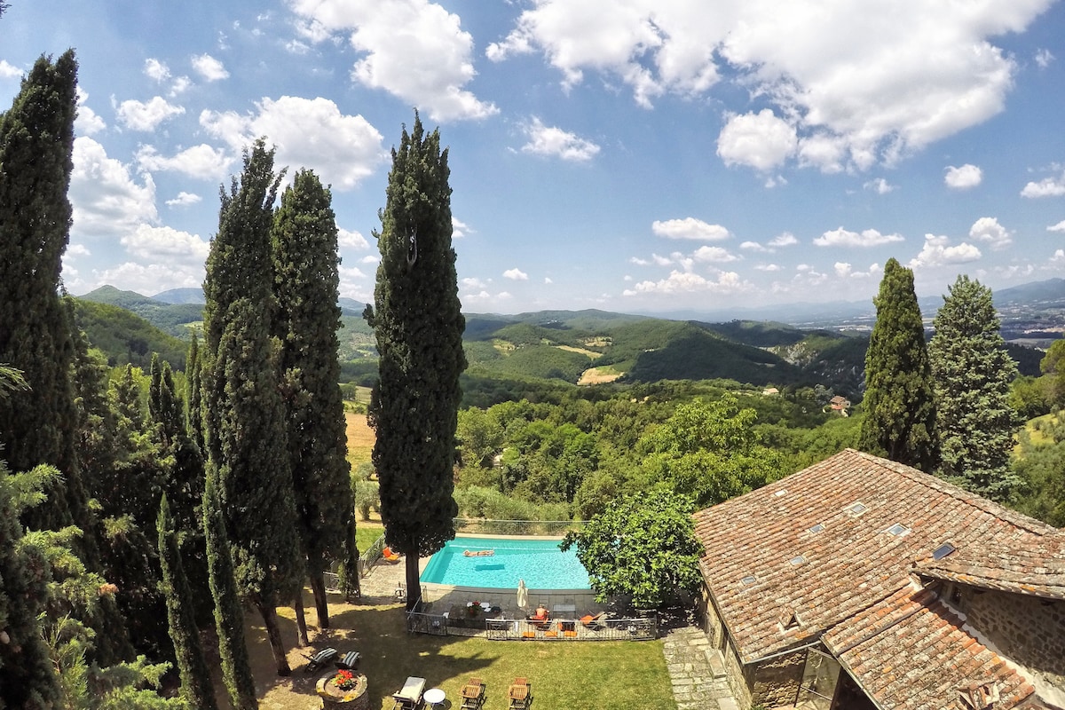 A panoramic view captures the rolling hills of the Mugello Valley, framed by tall cypress trees. A shared swimming pool is visible, surrounded by a well-kept garden and lounge chairs, creating a serene outdoor space for relaxation.