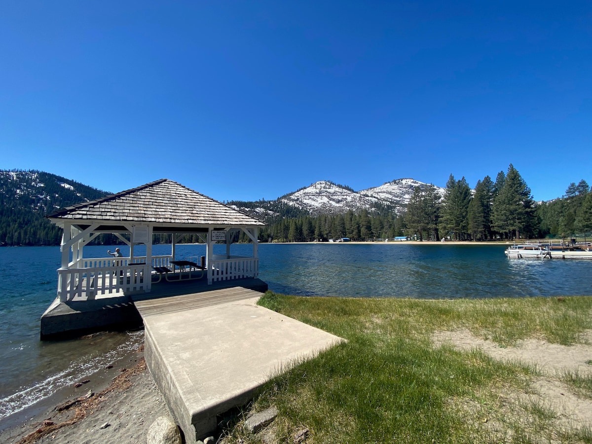 A serene lakeside view shows a wooden gazebo with a shingled roof, positioned on a concrete platform extending into the water. The surrounding landscape features grassy areas, pine trees, and snow-capped mountains in the background under a clear blue sky.