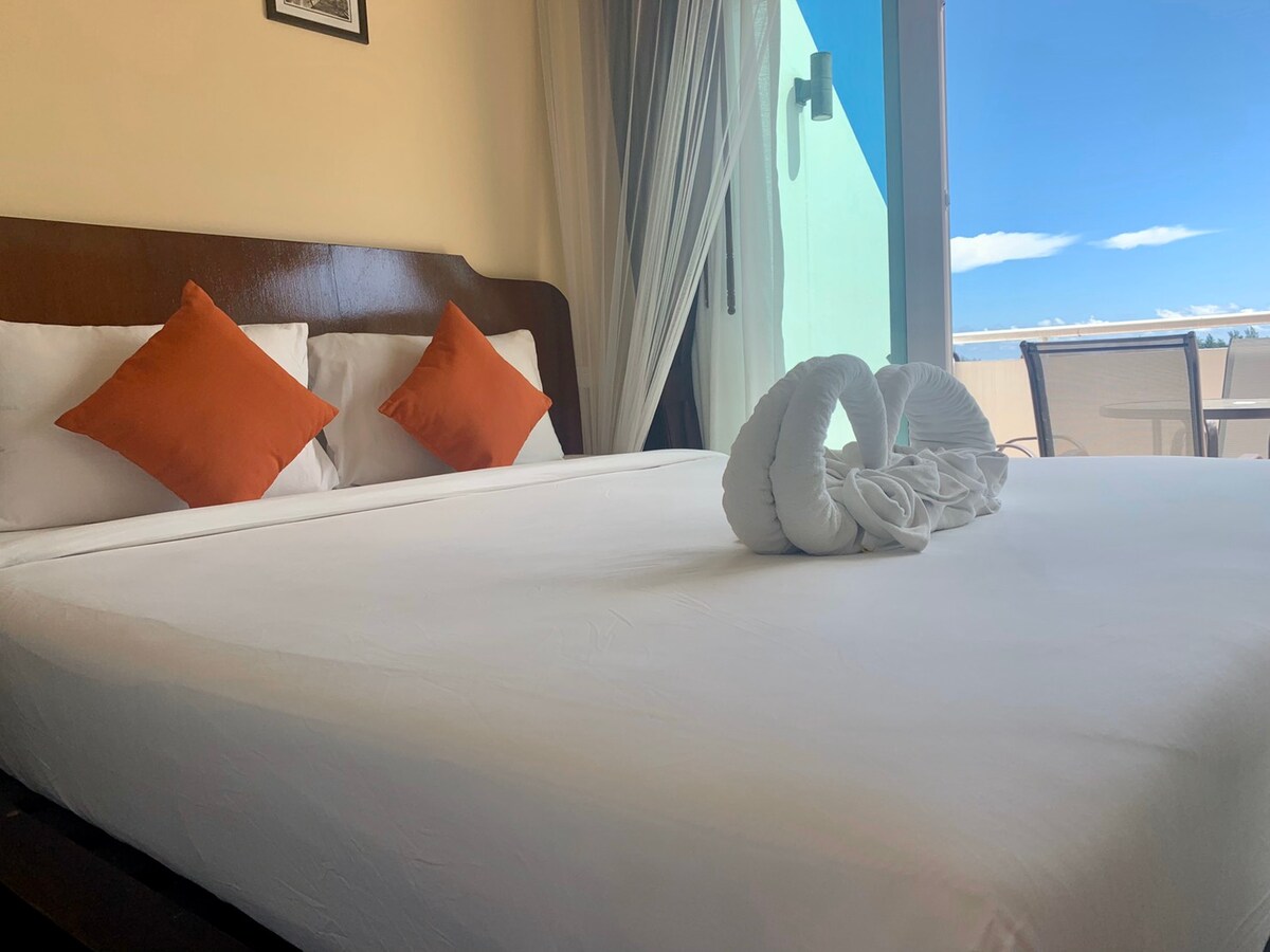 A spacious bed with crisp white linens is adorned with two orange accent pillows. A towel sculpture is elegantly placed at the foot of the bed. The floor-to-ceiling window offers a glimpse of the outdoor balcony and bright blue sky.