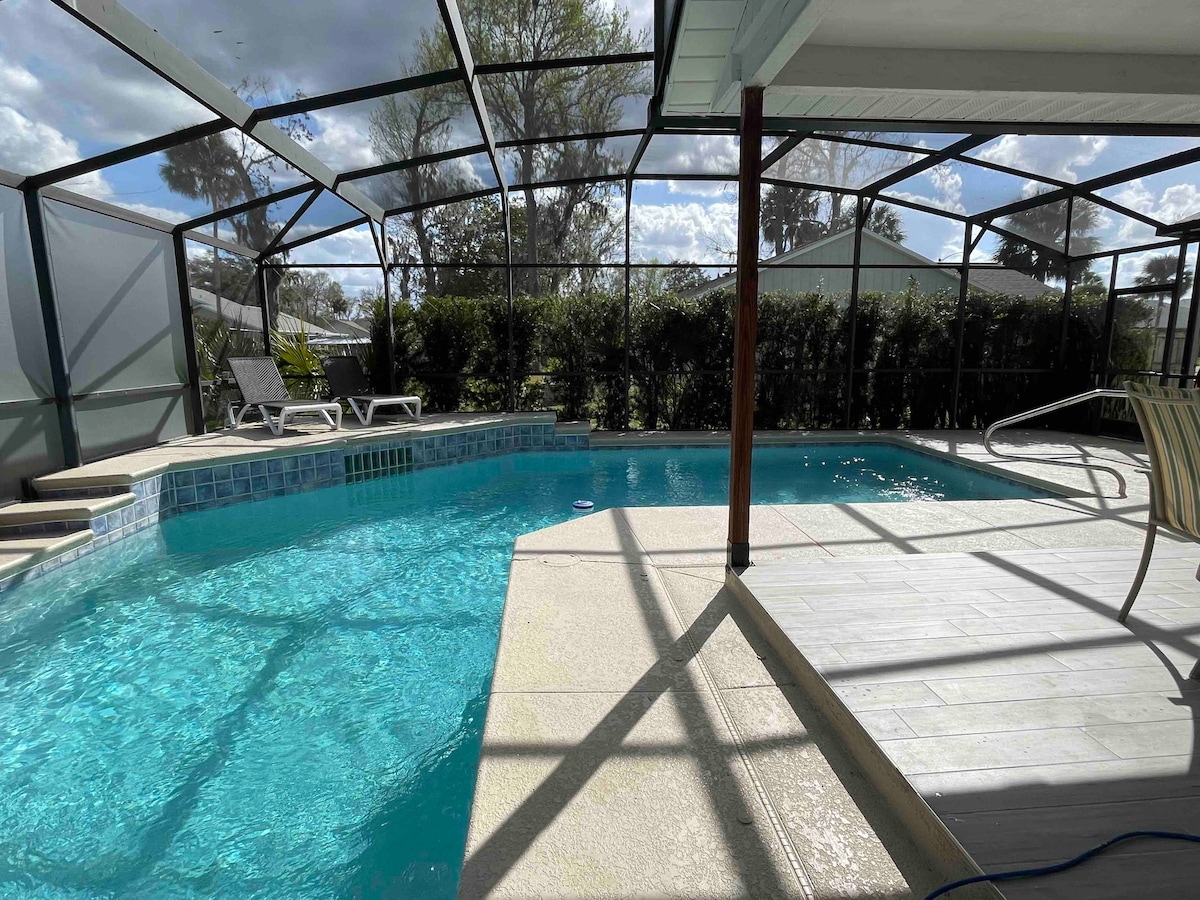 A private pool area features a clear blue pool surrounded by a screened enclosure. Two lounge chairs are positioned beside the water, and sunlight streams through the enclosure, creating a serene atmosphere. Lush greenery lines the perimeter, enhancing the tranquil setting.