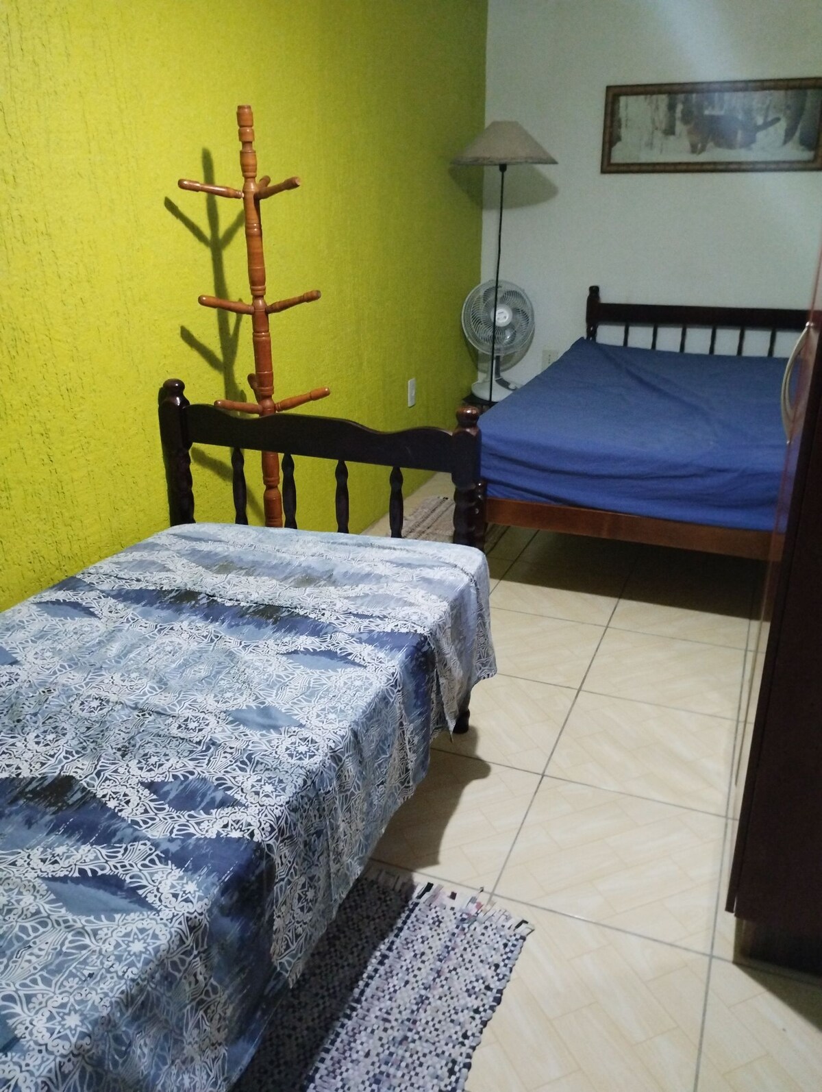 The room features a spacious double bed alongside a single bed draped in a patterned cover. A wooden coat rack stands near the beds, with a fan positioned for comfort. Green walls offer a bright backdrop, enhancing the room's inviting atmosphere.