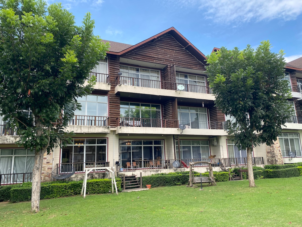 A multi-level building featuring wooden accents and large windows. Balconies are visible on each floor, framed by two small trees. The surrounding green lawn is well-maintained, with chairs arranged outside, creating an inviting outdoor space.