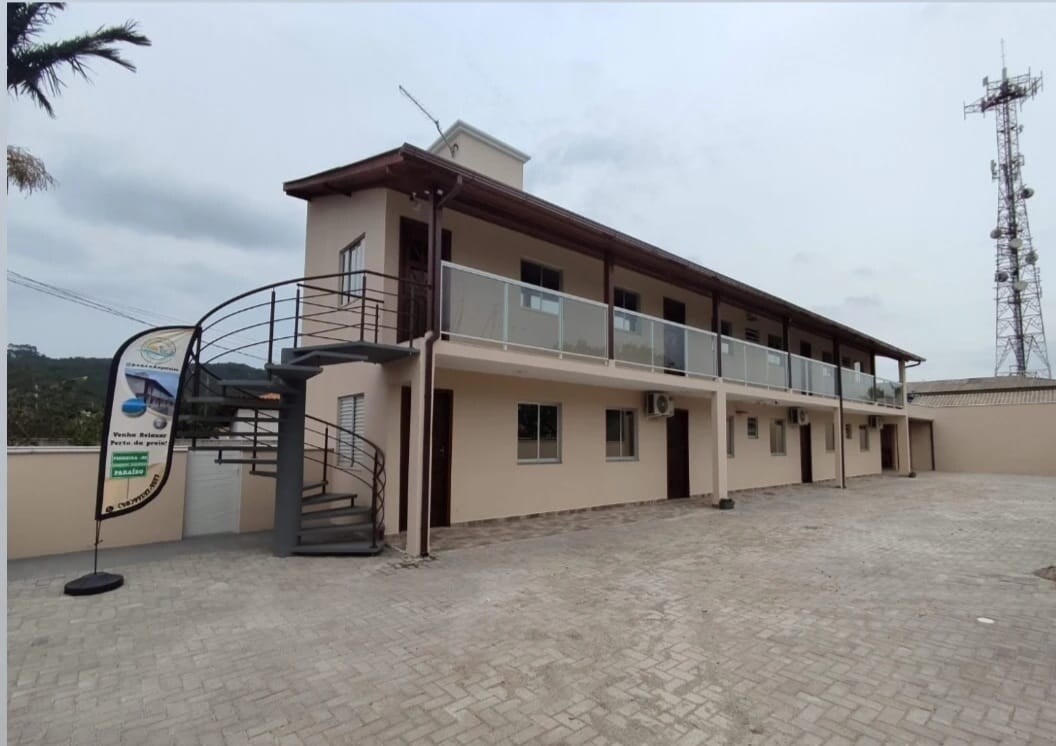 The exterior of a multi-story building features a modern design with a spiral staircase leading to a second-floor balcony. Large windows can be seen along the facade, and a telecommunications tower stands in the background. The surrounding area is paved with light-colored bricks.
