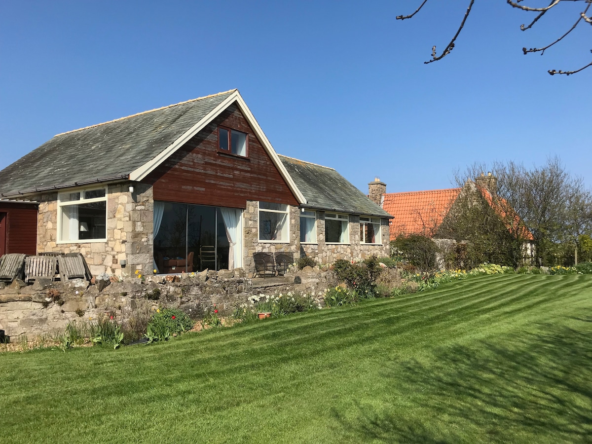 The spacious bungalow features a stone and wood exterior, blending harmoniously with the lush green lawn. Large windows allow natural light to fill the interior while offering views of the landscaped garden and surrounding scenery, providing a serene and open atmosphere.