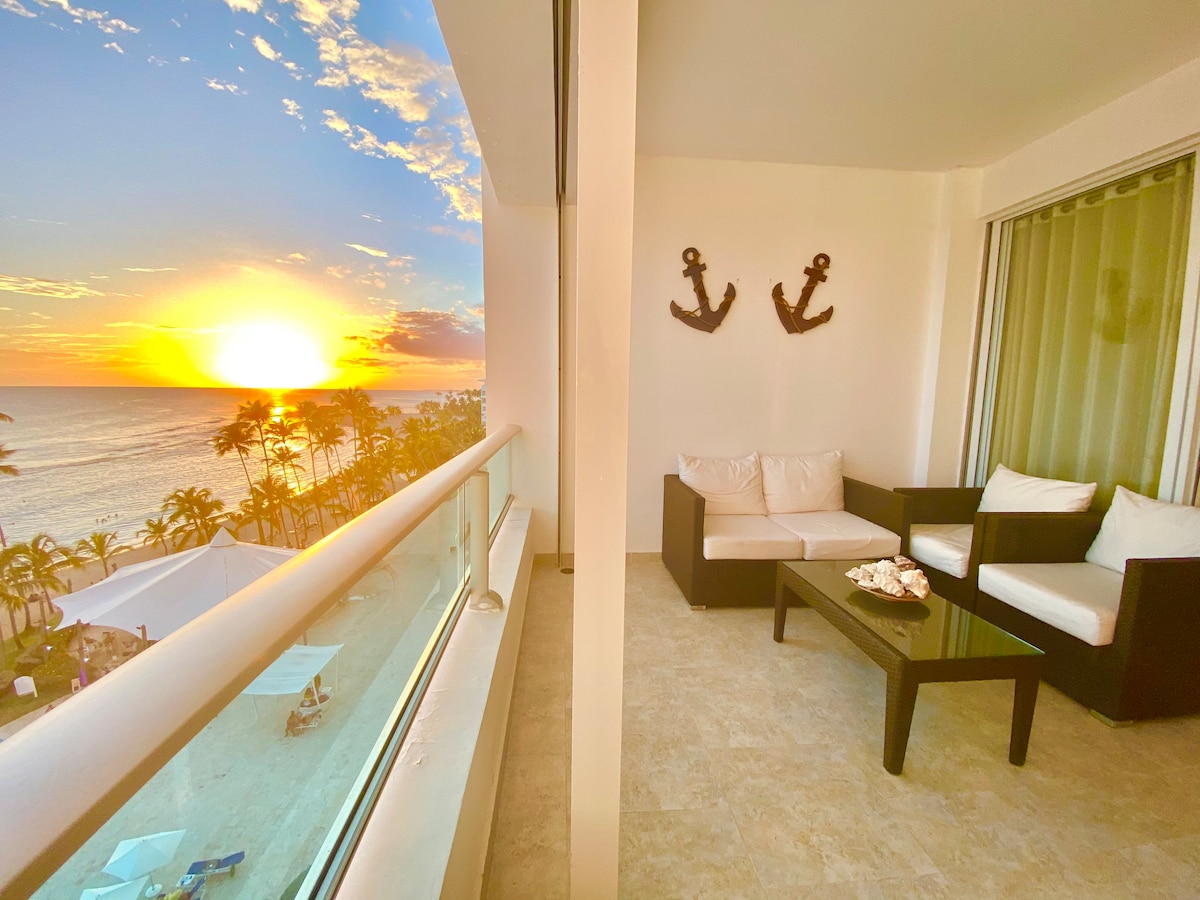 A balcony with a modern seating area overlooks a stunning sunset over the ocean. The elegant white sofa contrasts with dark wood accents, while two decorative anchors adorn the wall. Soft light reflects off the ocean, enhancing the serene atmosphere.