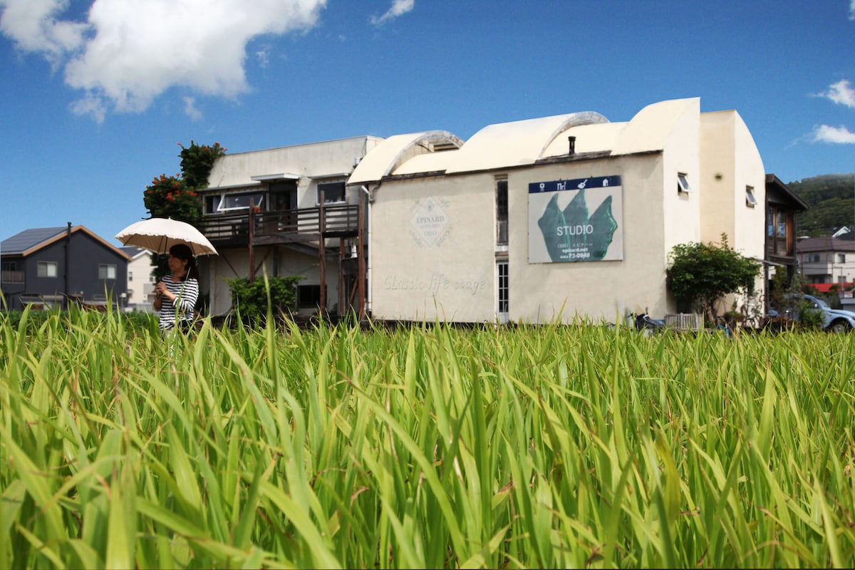 A charming studio building is situated amidst a lush green rice field, with a clear blue sky and scattered clouds above. The distinct architecture features unique curved roofing, while a person holding an umbrella walks through the tall grass, enhancing the serene countryside atmosphere.