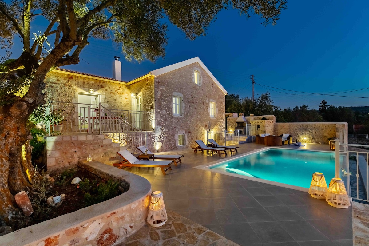 A traditional stone villa is illuminated at twilight, featuring a private pool surrounded by a spacious terrace. Sun loungers are positioned near the water, and lush olive trees provide natural shade. Soft lighting from the exterior highlights the villa's charming design and creates a serene atmosphere.