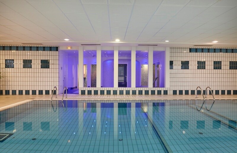 The indoor swimming pool is framed by tiled walls with a calming blue hue. Ambient lighting reflects off the water's surface, creating a serene atmosphere. Steps lead into the pool, while a doorway can be seen in the background, leading to additional spa facilities.