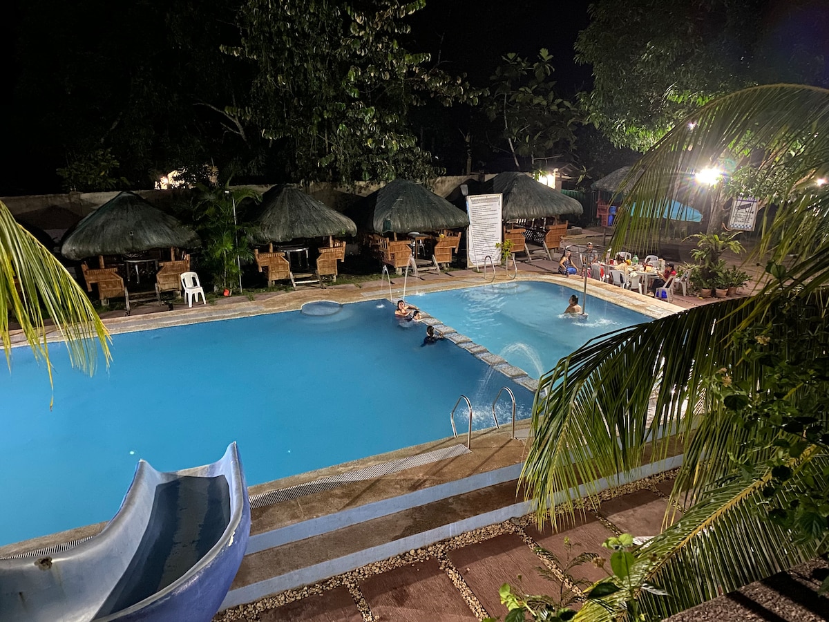 A swimming pool is seen under evening lights, featuring sparkling blue water. Lounge chairs are arranged around the pool's edge, with shaded areas provided by thatched umbrellas. A slide is positioned at one side of the pool, adding a playful element to the scene.
