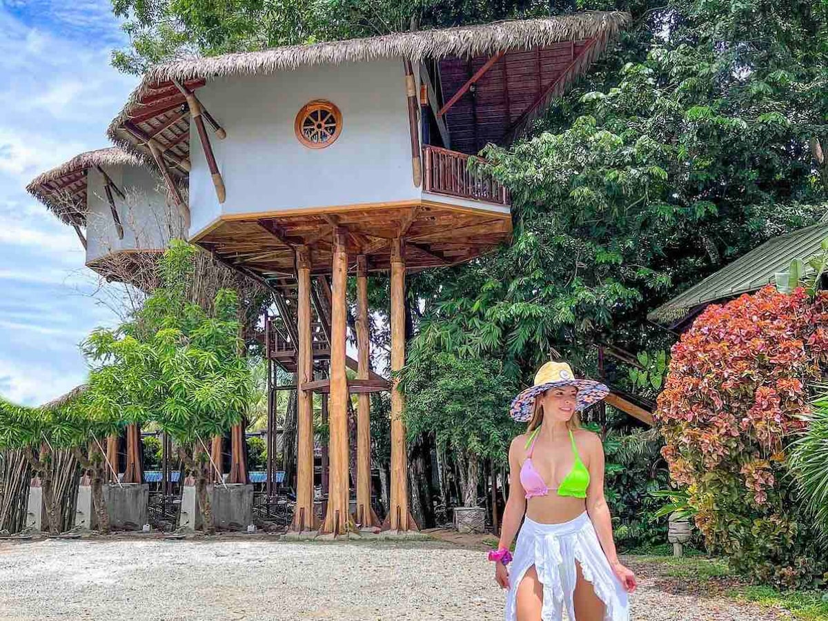 A unique treehouse structure stands elevated on sturdy wooden stilts, surrounded by lush greenery. The exterior features a thatched roof and round window accents. A guest is seen in a casual outfit, adding a sense of scale to the impressive architecture and natural surroundings.