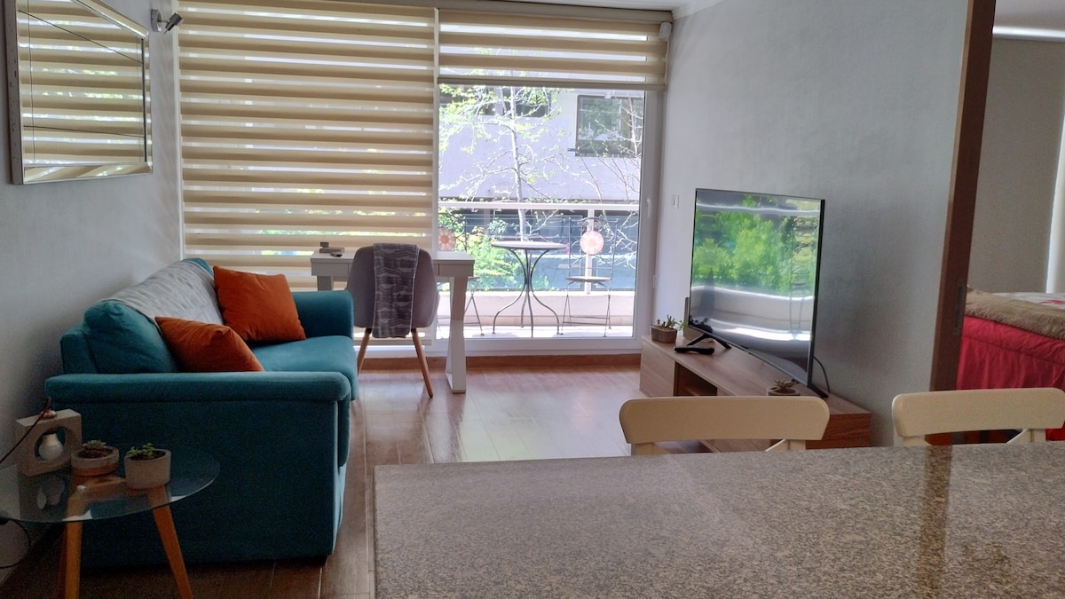 A modern living area features a turquoise sofa with orange cushions and a small glass table. A dining table can be seen in the background, while large windows with light-filtering blinds allow natural light to illuminate the space. A television is positioned on a low wooden console.