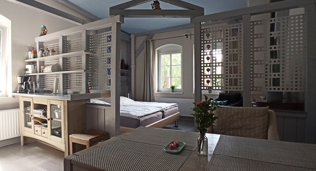 An open-concept room is divided by wooden structures, with a spacious bed and a small dining area featuring a table and chairs. A kitchenette, equipped with cabinets and essentials, is visible in the background. Natural light enters through a window, highlighting the neutral color scheme.
