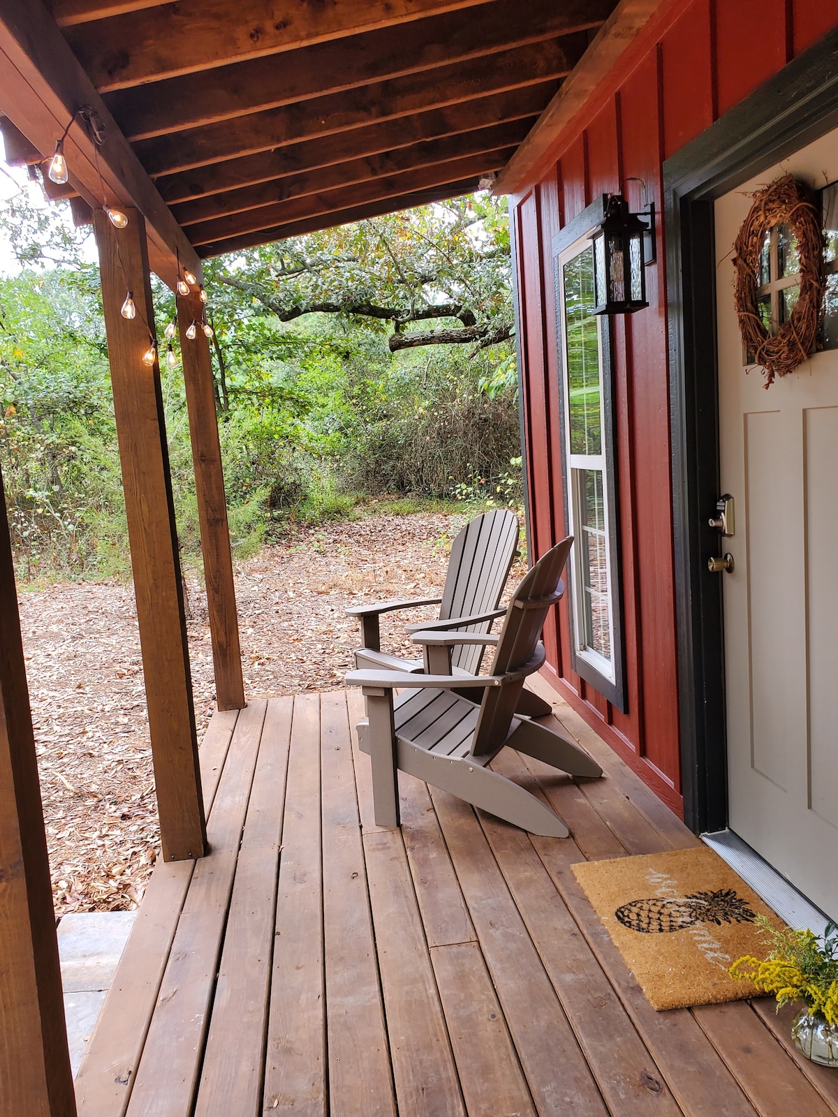 A covered front porch is shown, featuring two Adirondack chairs positioned on wooden flooring. Soft outdoor lighting is visible, while a welcoming doormat is placed near the entrance. Lush greenery surrounds the area, enhancing the serene, secluded ambiance.