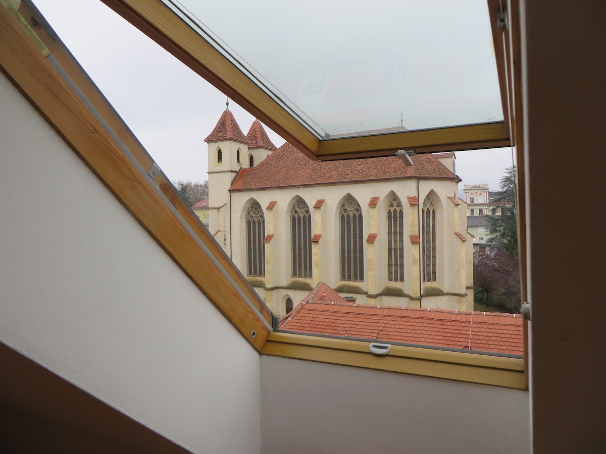 A view from a skylight framed by wooden beams reveals a charming church with tall, arched windows and a red-tiled roof. The building's unique architectural features are visible against a backdrop of a gray sky.