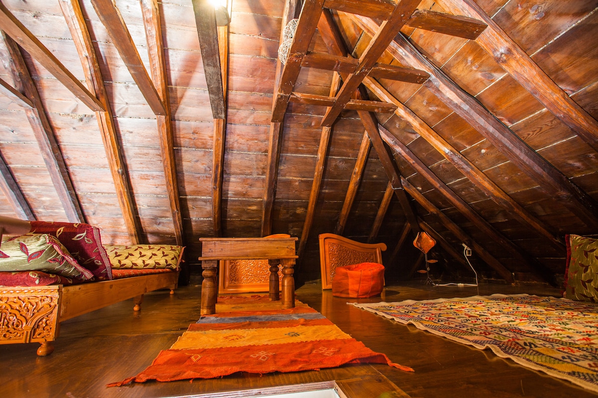 The attic space features a striking wooden ceiling with exposed beams, enhancing the room's character. Various seating options are arranged around a central wooden table, with colorful textiles adorning the floor. Natural light filters in, creating a warm and inviting atmosphere.