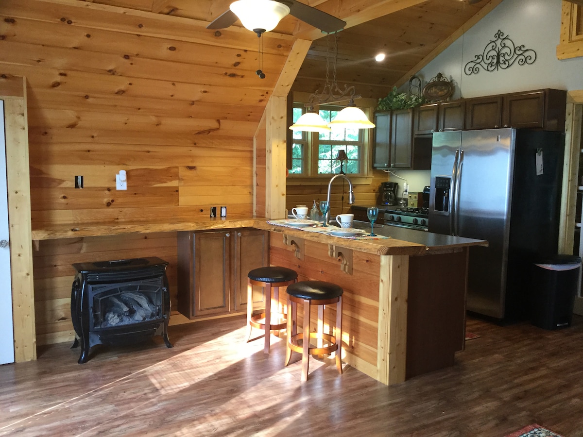 An inviting kitchen area features wooden cabinetry and a spacious island with two stools. A gas log stove is positioned near the entrance, while natural light pours in from large windows above the sink, highlighting the warm wood paneling throughout the space.