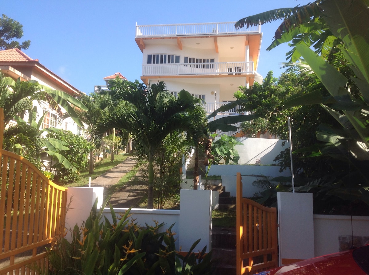 A two-story property is framed by vibrant greenery, with a pathway leading to its entrance. The building features a balcony on the upper level, surrounded by tropical plants. A welcoming entrance gate is positioned in the foreground, inviting guests into the lush landscape.