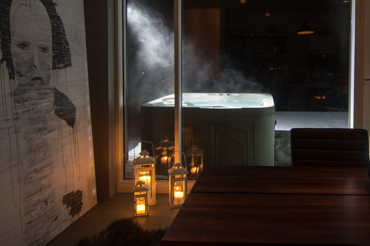 A modern indoor space features a large window revealing a steaming hot tub outside. Soft light from lanterns on the floor creates a warm ambiance, while a minimalist artwork hangs beside a sturdy dining table. The space blends contemporary design with relaxation elements.