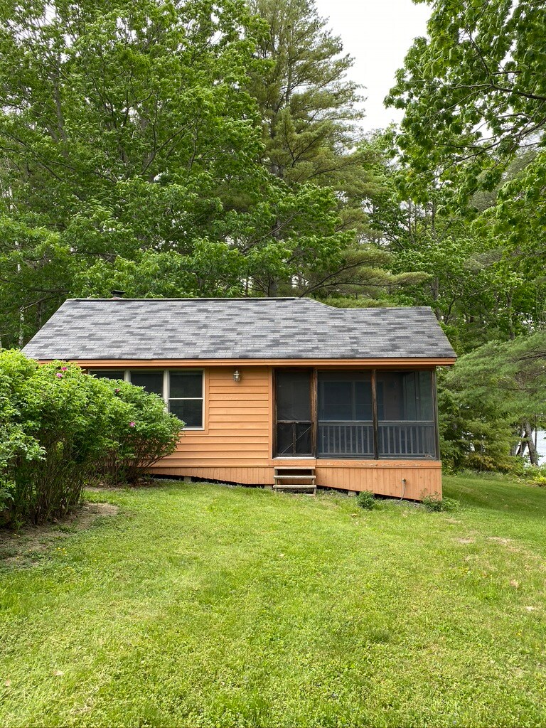 A cozy cabin is nestled among lush green trees, showcasing an inviting screened-in porch. The exterior features warm orange siding and a sloped roof, with stepping stones leading up to the entrance. Surrounding foliage adds to the cabin's secluded charm.