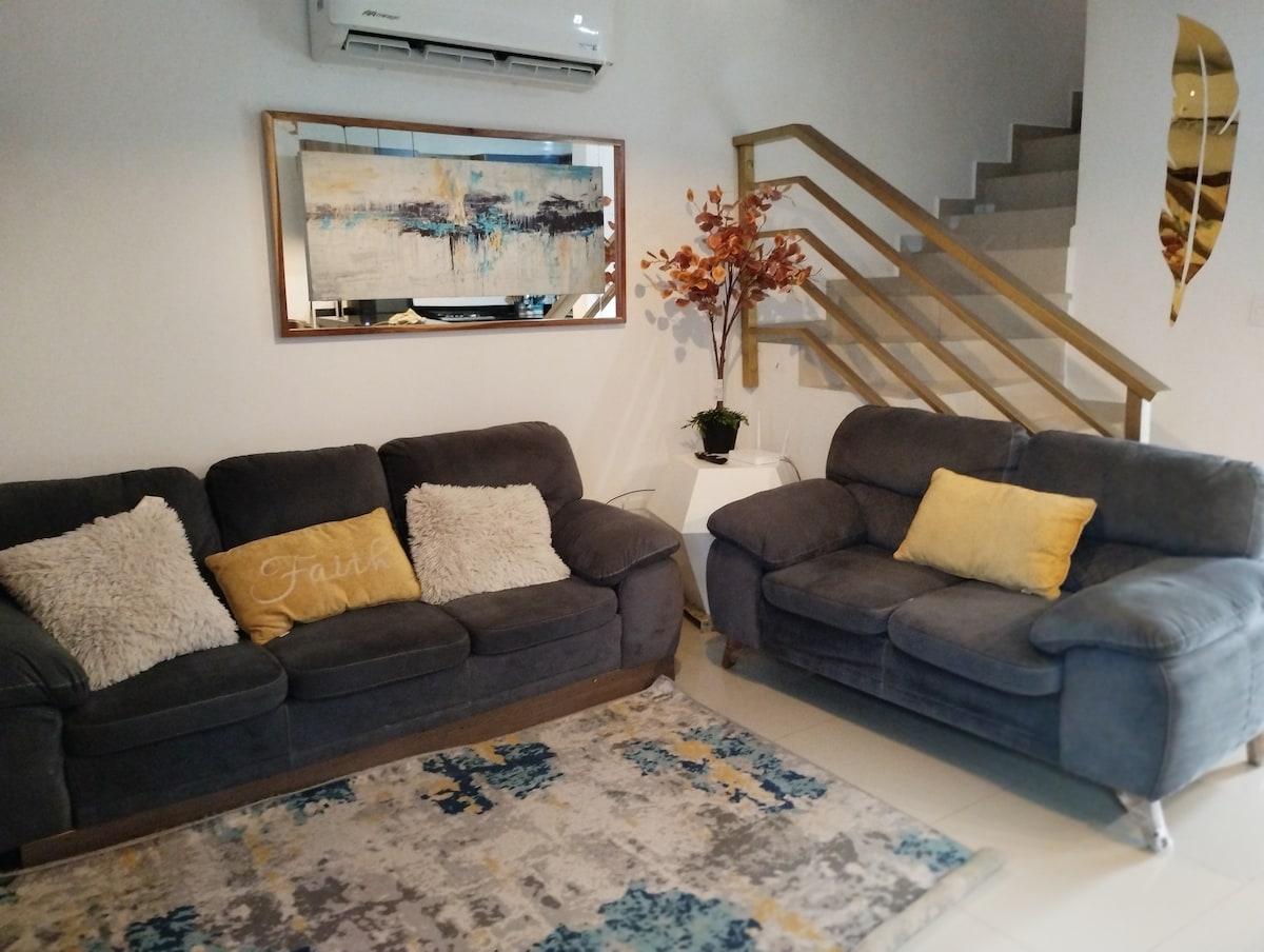 A comfortable living area features two grey sofas with textured cushions and a decorative yellow pillow. A decorative rug adds color to the space. A potted plant and a piece of wall art are present, while a staircase is visible in the background.