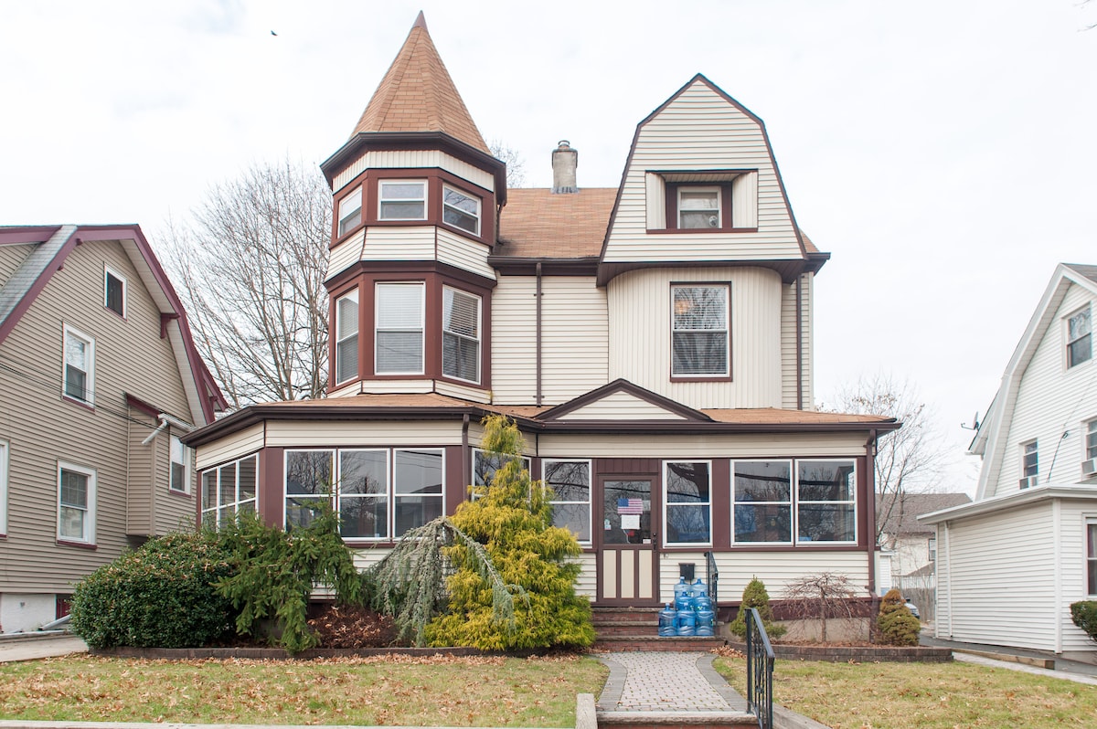 The exterior of a Victorian-style house is shown, featuring a pointed turret and large windows framed by greenery. A pathway leads to the entrance, with a well-maintained lawn that includes bushes and a small tree, creating a welcoming ambiance.