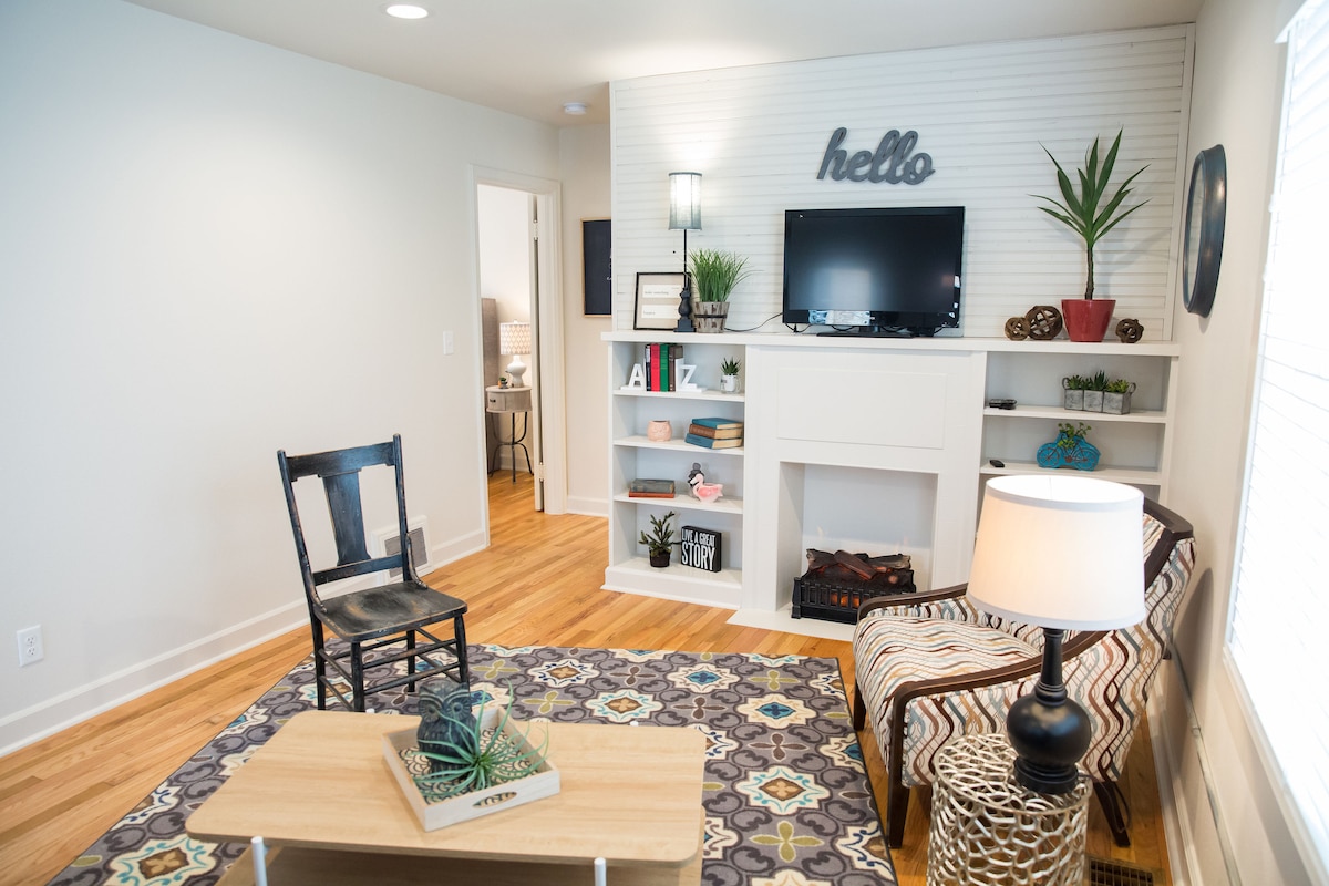 The living room is characterized by a stylish arrangement, featuring a plush sofa and a wooden coffee table. A wall-mounted TV is positioned above an electric fireplace, flanked by decorative shelves. A cozy patterned rug and a chair enhance the inviting atmosphere.